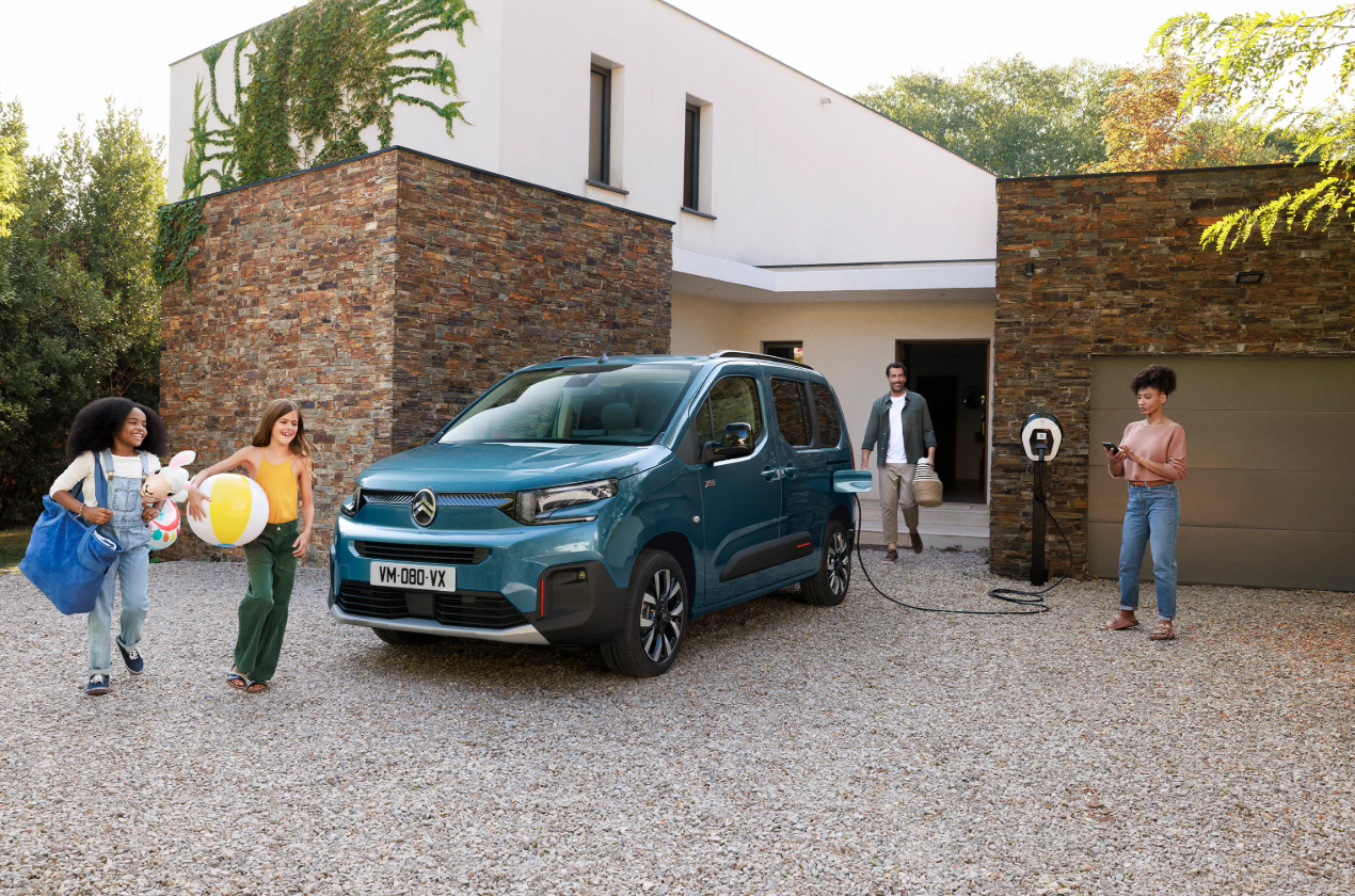 Voorzijde van de Citroën ë-Berlingo, aangesloten op een laadpaal, geparkeerd voor een huis met mensen die naar de auto lopen.